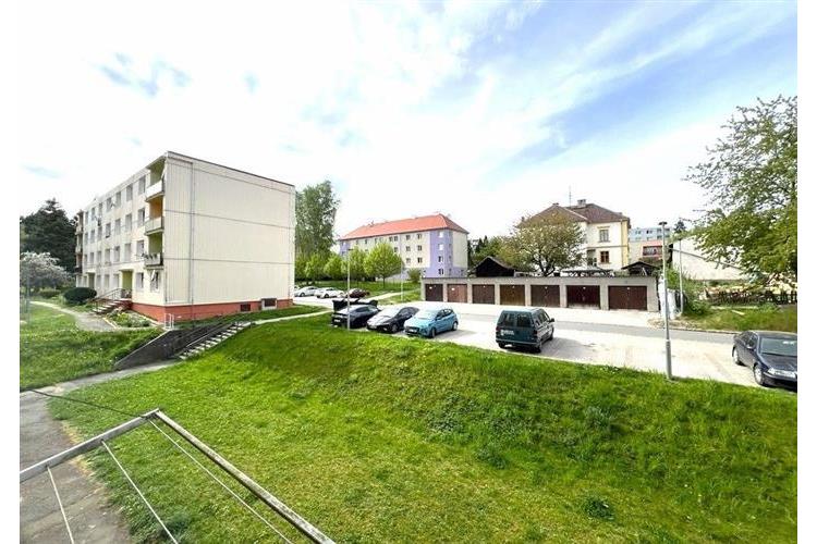 Výhled na moderní venkovní parkoviště s několika auty, obklopené zelení a bytovými domy, dobrá lokalita pro bydlení.