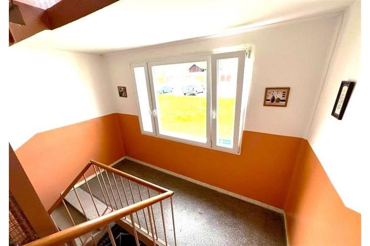 Světlé schodiště s velkým oknem, dlažba na podlaze a moderní zábradlí. Pečlivě udržovaný prostor s přirozeným světlem.