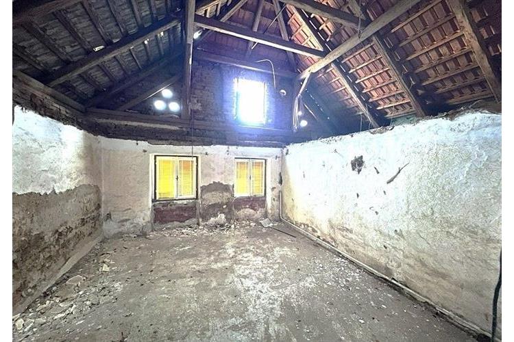 Podkrovní prostor s vysokým stropem, dřevěné trámy, nutná rekonstrukce, přirozené světlo skrz malá okna, neomítnuté cihlové zdi.