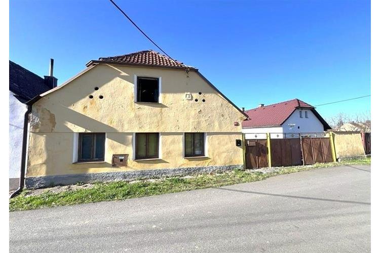 Exteriér venkovského domu s dřevěnými okenicemi a sedlovou střechou, vhodný pro rekonstrukci, nachází se v klidné lokalitě s výhledem na ulici.