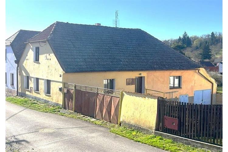 Kamenný venkovský dům s tradiční šikmou střechou a dřevěnými okenicemi se nachází na klidné ulici v malebné krajině.