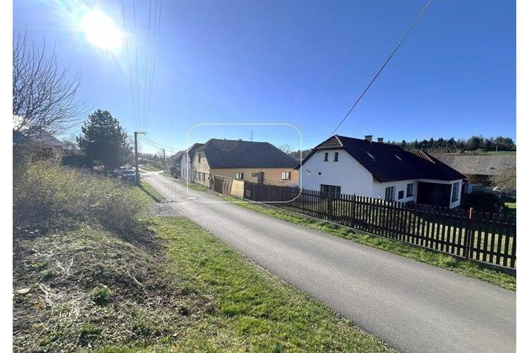 Klidná venkovská ulice s rodinnými domy a zahradami, asfaltová silnice, slunný den, přírodní okolí, ideální pro bydlení mimo město