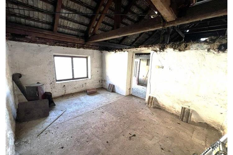 Podkrovní místnost s dřevěnými trámy a betonovou podlahou, vhodná k rekonstrukci, má velké okno poskytující dostatek přirozeného světla.